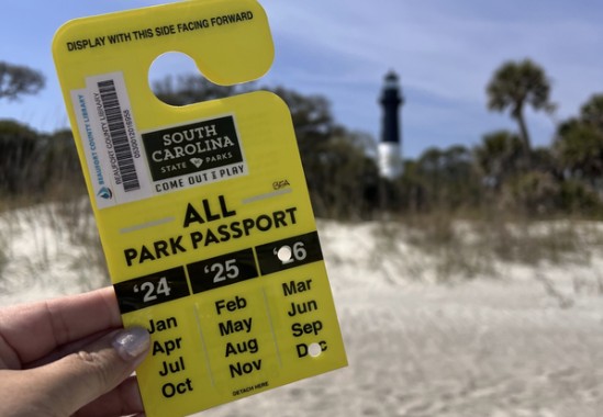 Close-up of hand holding SC State Park Pass in the foreground, with the Hunting Island Lighthouse and palm tree blurred in the background.